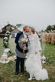 Newlyweds stand near wedding arch and kiss next to dog photo
