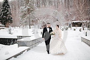 Newlyweds in witer park are walking around. Handsome groom and beautiful bride surrounded by snow. Winter wedding