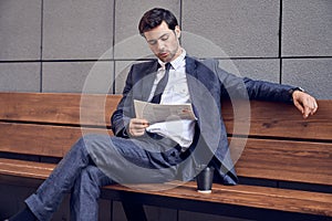 Daily news. Full length of a pleasant smiling businessman reading a newspaper with coffee while sitting on the bench