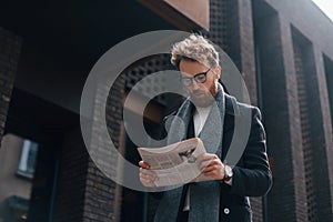 With newspaper in hands. Stylish man with beard and in glasses is outdoors near building
