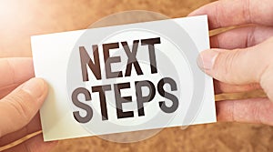 NEXT STEPS word inscription on white card paper sheet in hands of a businessman. recap concept. red and white paper