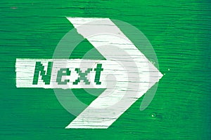 `Next` text written on a white directional arrow painted on a green wooden signboard background.