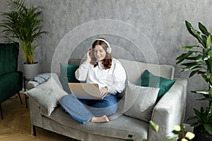 Nice beautiful 30s woman with headphones working at the laptop and sitting on the sofa at home. Concepts of education