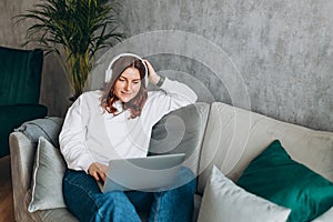 Nice beautiful 30s woman with headphones working at the laptop and sitting on the sofa at home. Concepts of education