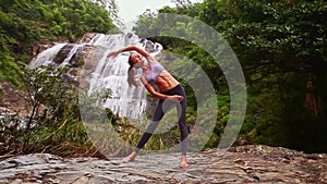 Nice blond girl bends body in yoga pose by waterfall