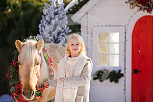 Nice blonde curly child caresses adorable pony with festive wreath near the small wooden house and snow-covered trees. New Year an