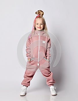 Nice blonde kid girl with girlish hairstyle and colorful braids in pink jumpsuit stands with hands in her pockets