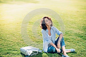 Nice brunette girl with short hair is chilling on grass in park . She wears white T-shirt, shirt and jeans, shoes. She