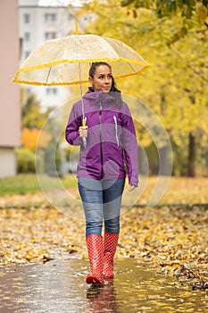 A nice brunette in warm clothes and with an umbrella is walking in a city park surrounded by fallen autumn leaves