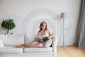 Nice brunette woman in pink clothes reading a magazine and sitting on sofa on white studio background