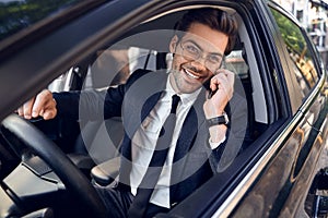 Nice business talk. Handsome young businessman talking on his smart phone and smiling while sitting on the front seat