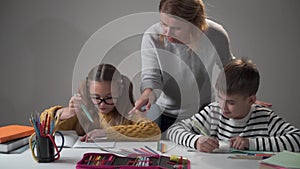 Nice Caucasian mother helping her children doing homework. Little cute girl in eyeglasses and her brother sitting at the