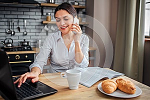 Nice cheerful woman sit at table and work at home. She typing on laptop keyboard and talk on phone. Her breakfast stand