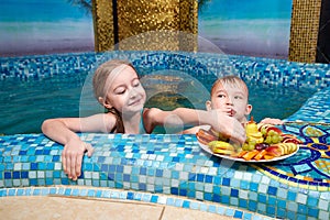 Nice children in blue small pool with plate of fruits. Girl and boy have fun in water indoors