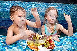 Nice children in blue small pool with plate of fruits. Girl and boy have fun in water indoors