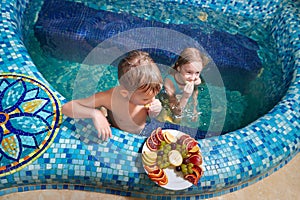 Nice children in blue small pool with plate of fruits. Girl and boy have fun in water indoors