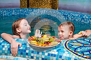 Nice children in blue small pool with plate of fruits. Girl and boy have fun in water indoors