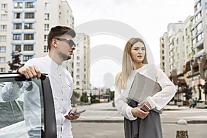 Nice couple are standing together outside on street. Guy is leaning on car`s door. He wears glasses. Girl is looking at the same