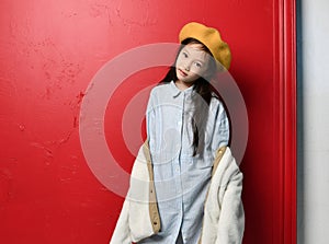 Nice cute asian kid girl iblue shirtdress felt beret and beige jacket is posing over red