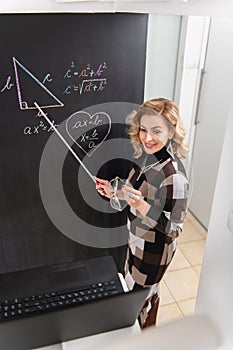 Nice dressed young female math teacher explaining geometry on the black wall. Online working from home. Covid 19 pandemic