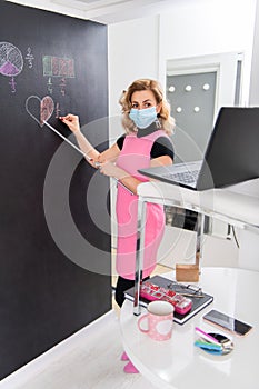 Nice dressed young female math teacher wearing mask and explaining fractals on the black wall. Online working from home. Covid 19
