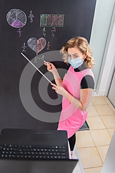 Nice dressed young female math teacher wearing mask and explaining fractals on the black wall. Online working from home. Covid 19