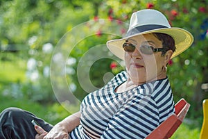 Nice elderly lady in a hat and sunglasses sitting on a chair in the garden