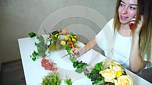 Nice female flower designer accepts order for flower arrangement on phone, sitting at table in bright office in daytime