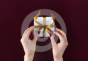 Nice female hands holding gift box wrapped in white paper with golden ribbon on festive background
