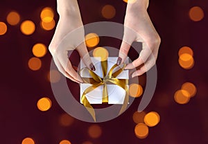 Nice female hands holding gift box wrapped in white paper with golden ribbon on festive background