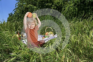 nice and funny fair-haired kid holds an apple and orange, basket with fruits on green grass