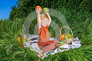 nice and funny fair-haired kid holds an orange over his head, basket with fruits on green grass