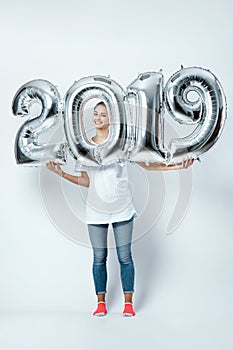 Nice funny girl dressed in white t-shirt, jeans and pink socks holding balloons in the shape of numbers 2019 on the