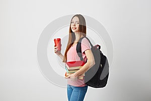 Nice girl with a black backpack on her shoulder dressed in a pink t-shirt and jeans holds a red plastic cup and books in