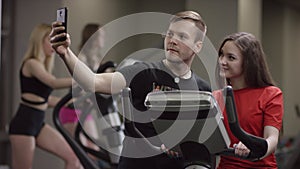 Nice girl is diligently working on an exercise bike and cool guy come to her and suggests to make selfie on a background