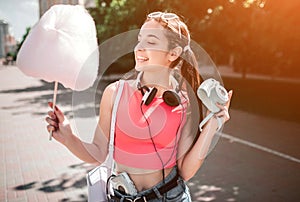 Nice girl is posing. She is holding cotton candy in one hand and camera in the other one. Also there is headphones