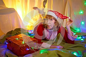 Nice girl in a red dress and hat of Santa with big red gift box in room decorated for Christmas. Woman in beautirul