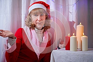 Nice girl in red dress and hat of Santa in room with candles before Christmas. Woman in beautirul studio during photo shoot before