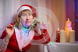 Nice girl in red dress and hat of Santa in room with candles before Christmas. Woman in beautirul studio during photo shoot before