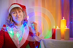 Nice girl in red dress and hat of Santa in room with candles before Christmas. Woman in beautirul studio during photo shoot before