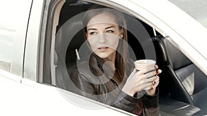 Nice girl sits in the front seat in the car and drinks coffee.