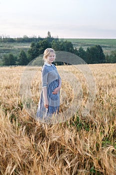 Nice happy young girl in blue old-fashioned outdoor dress in golden field of ears at dawn. Green boscage in background