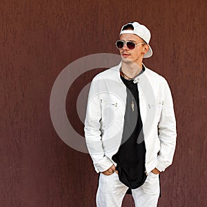 Nice hipster young man in a trendy white summer jacket in a stylish black t-shirt in a fashionable baseball cap in dark sunglasses