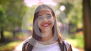 Nice lady is standing in park in daytime, watching at camera, smile, blurred backgrouns