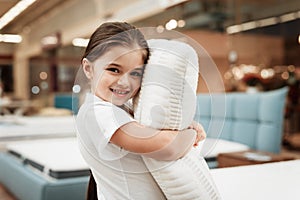 Nice little girl hugs pillow in store of orthopedic mattresses. Testing softness of pillow.