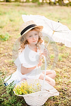 A nice little girl with light curls in a straw hat, a white dress and a lace umbrella with a basket of flowers