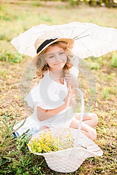 A nice little girl with light curls in a straw hat, a white dress and a lace umbrella with a basket of flowers