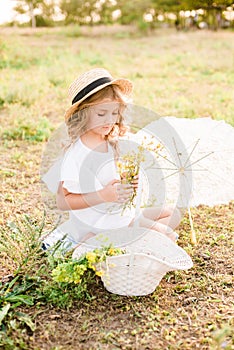 A nice little girl with light curls in a straw hat, a white dress and a lace umbrella with a basket of flowers