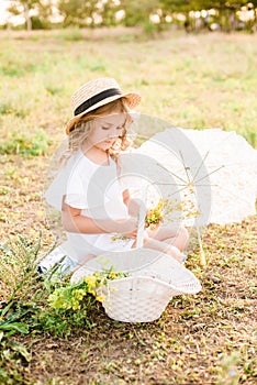 A nice little girl with light curls in a straw hat, a white dress and a lace umbrella with a basket of flowers