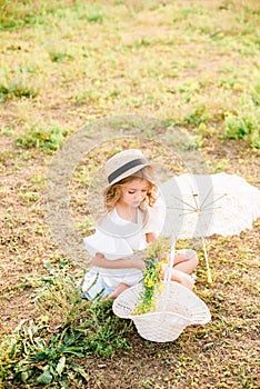 A nice little girl with light curls in a straw hat, a white dress and a lace umbrella with a basket of flowers